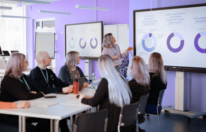 Ihmisiä luokkahuoneessa pöydän äärellä. Yksi esittelee seisaaltaan näytöllä olevia ympyrädiagrammeja.